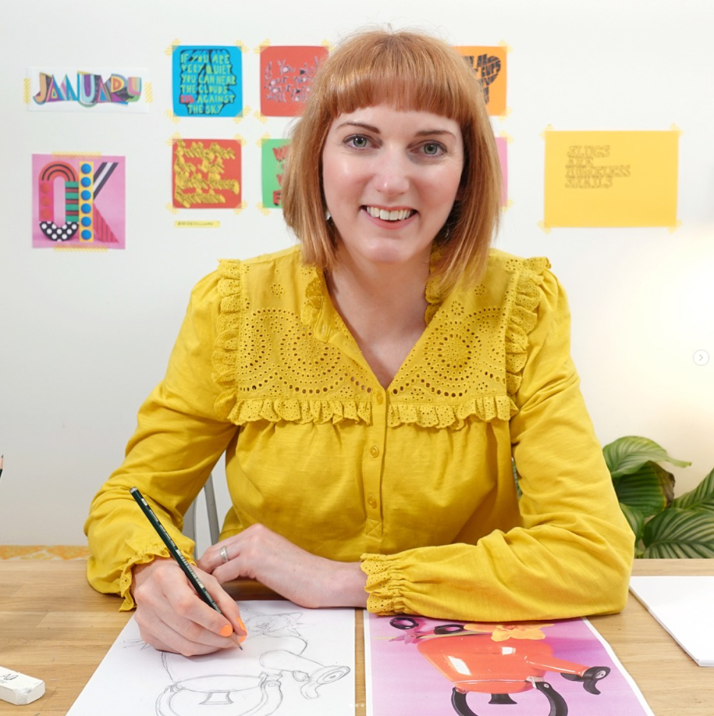 A person with chin length ginger hair with a fringe smiling into the camera, sat at a desk drawing illustrations.