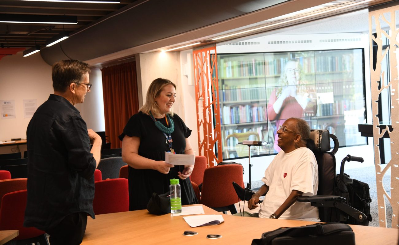 A white man, a white women and a black man in a wheelchair around a table laughing and talking together.