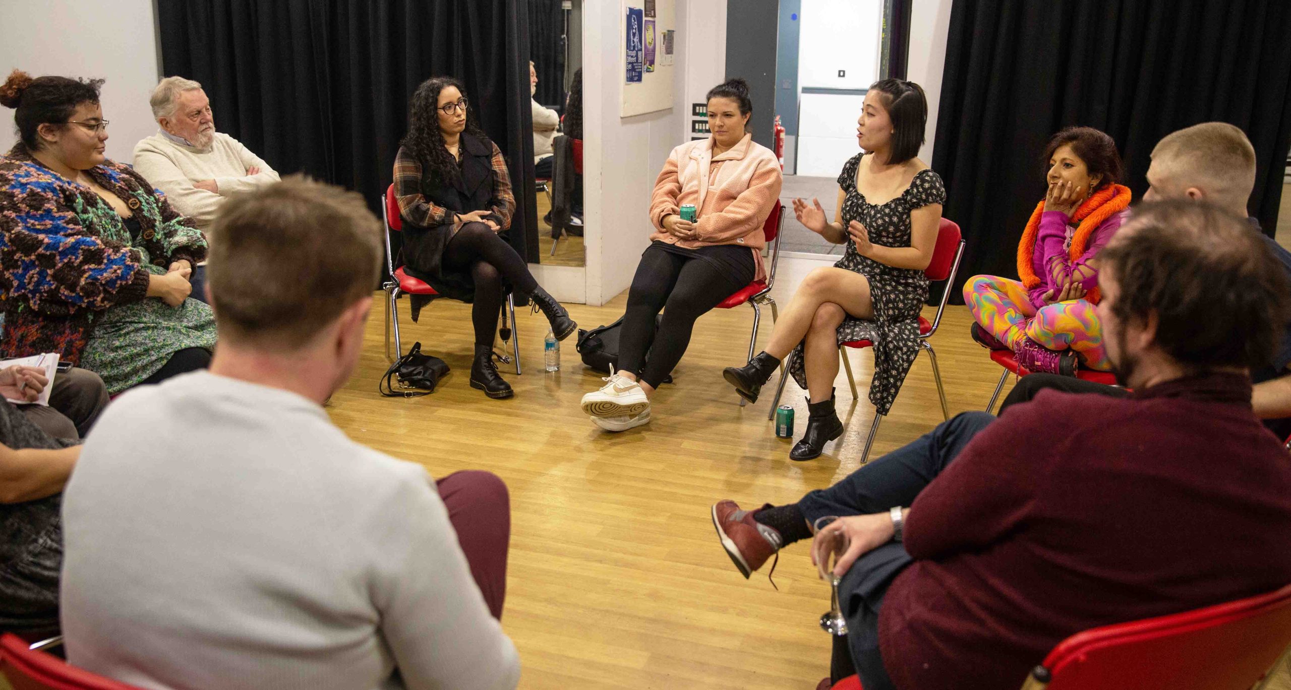 A group of people sat in chairs in a circle discussing different topics in a dance studio.