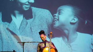 Celloist, Ayanna Witter-Johnson, holding a cello and looking down while smiling. In the background is a giant photo of a mother and daughter smiling at each other.