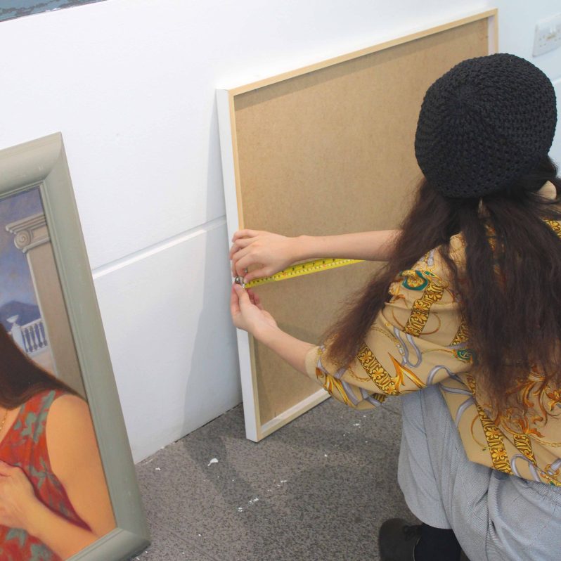 A person crouched down wearing a black beret with long hair, measuring a frame with a measuring tape.