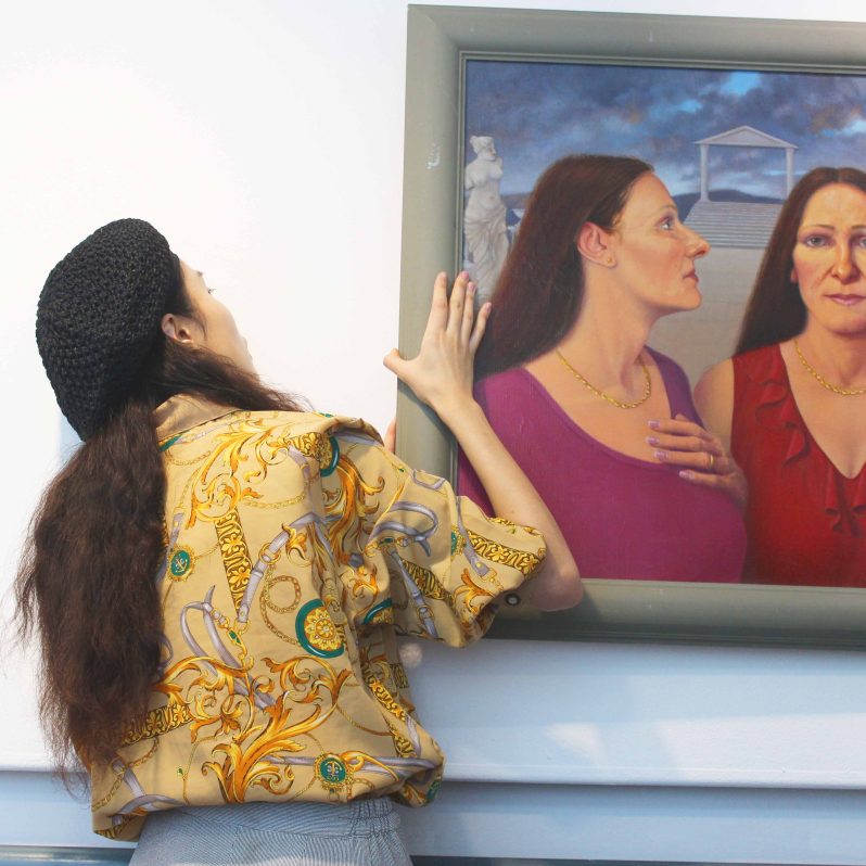 A women with long dark hair, a black beret and a yellow shirt straightening a painting on the wall with their hands.