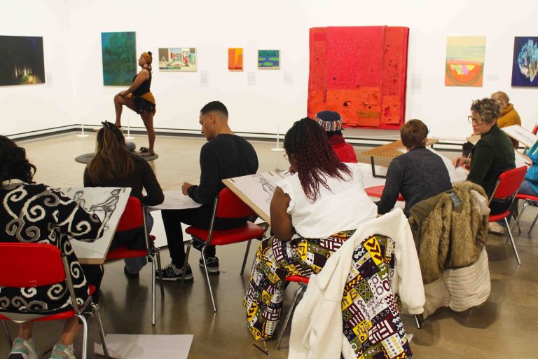 A group of people sat down with paper and pencils in a art gallery drawing a Black women posing in the background.