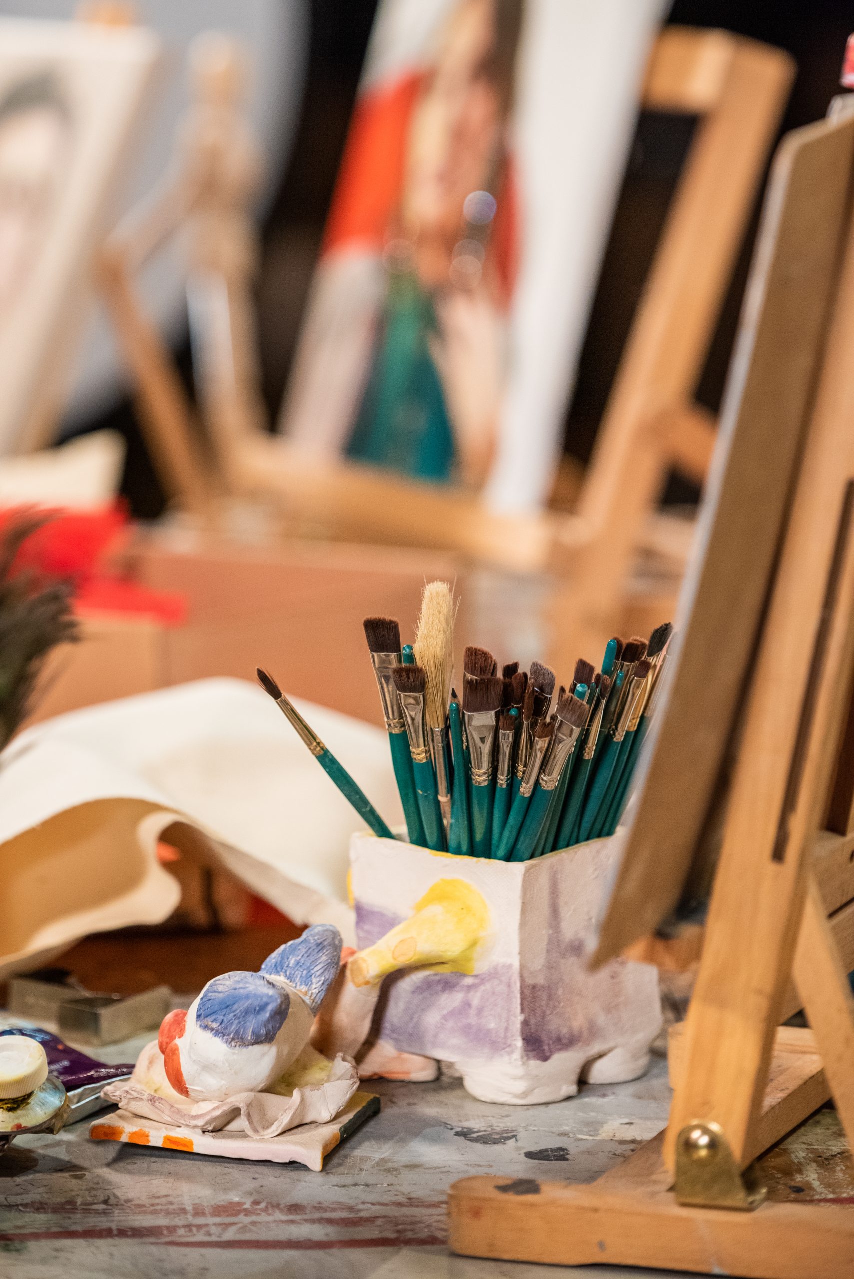 A messy paint studio with oil paintings in the background and paintbrushes in a pot in the foreground.