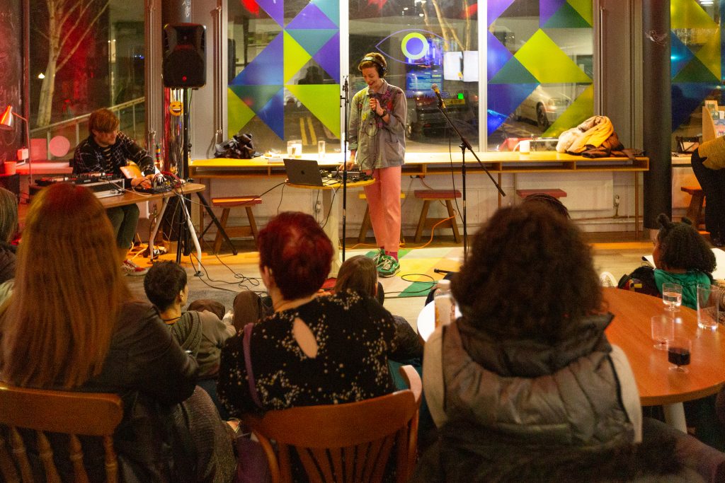 A women comedian performing with a microphone in the AAC cafe with a crowd.