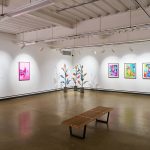 A large white gallery with colourful paintings on the wall and glass flower sculptures in the corner.