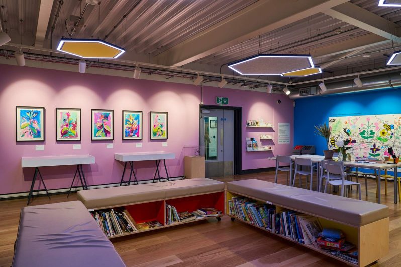 A colourful room with pink and blue walls, hanging artwork on them. In the middle of the room are tables and chairs.