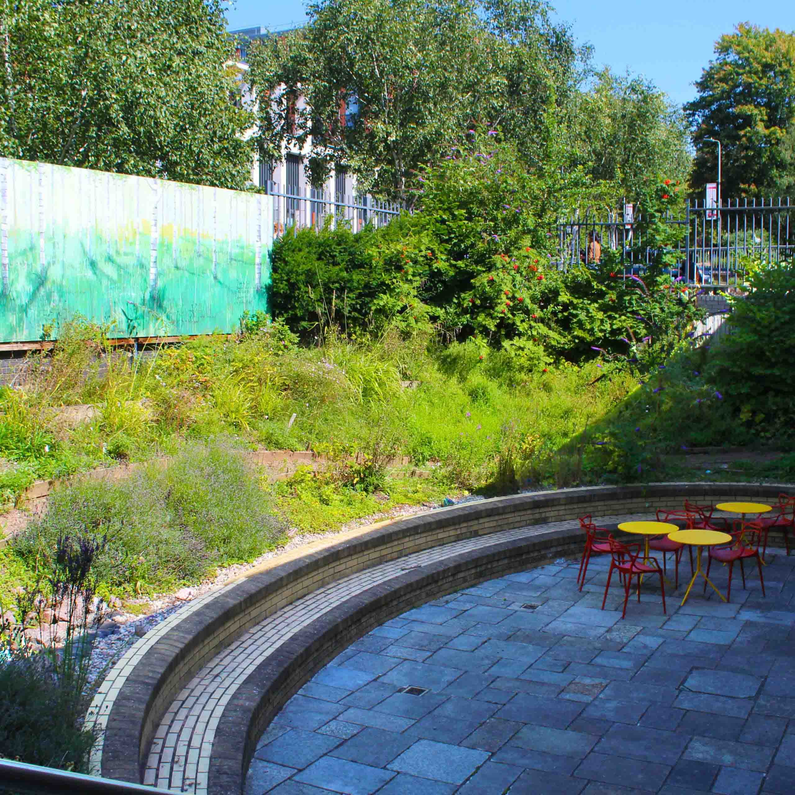 A semi-circle of lush greenery with a bricked area in the centre featuring tables and chairs.