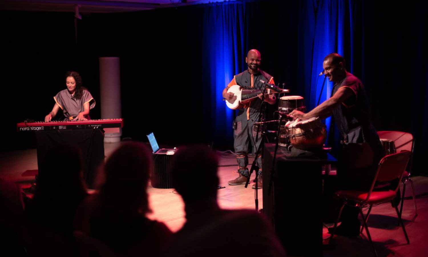 Three instrumentals are playing drums, a keyboard and a sitar, dressed in grey clothing with a crowd in the foreground.