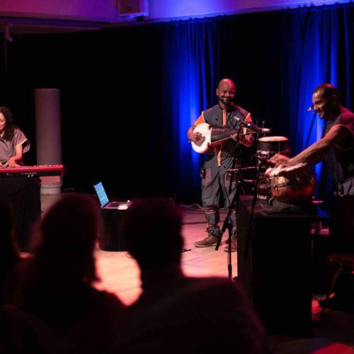 Three instrumentals are playing drums, a keyboard and a sitar, dressed in grey clothing with a crowd in the foreground.