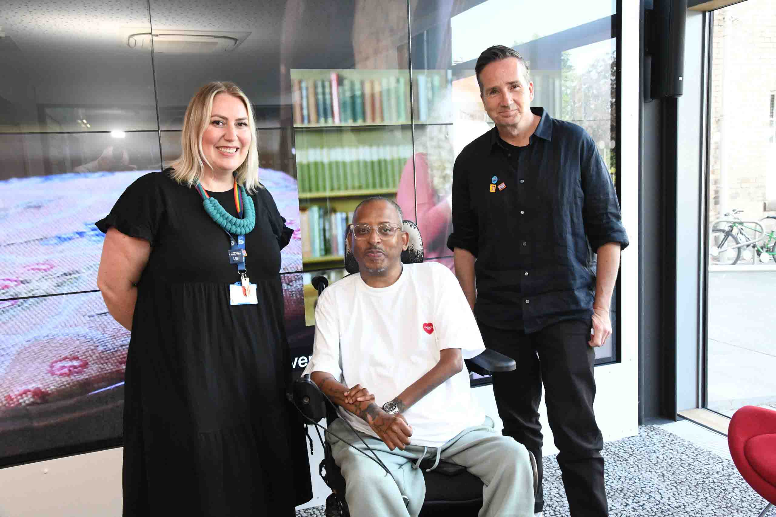 A women, a man in a wheelchair and another man standing in front of a black screen, smiling.