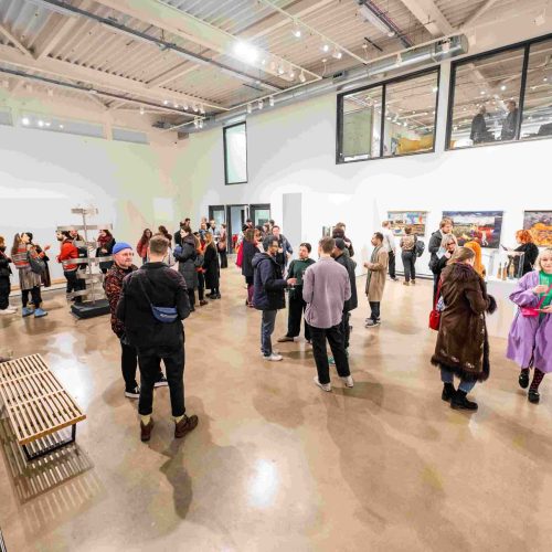 A large group of people wander around a large, white gallery space looking at the artwork.