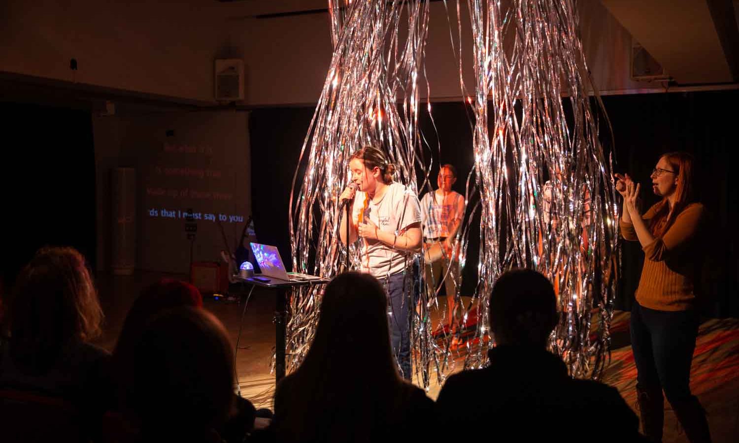 A women sings into a microphone while looking at a laptop. Behind her is glittering strings floating down from the ceiling with people blowing the strings behind her.