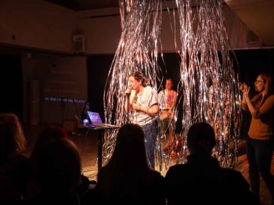 A women sings into a microphone while looking at a laptop. Behind her is glittering strings floating down from the ceiling with people blowing the strings behind her.