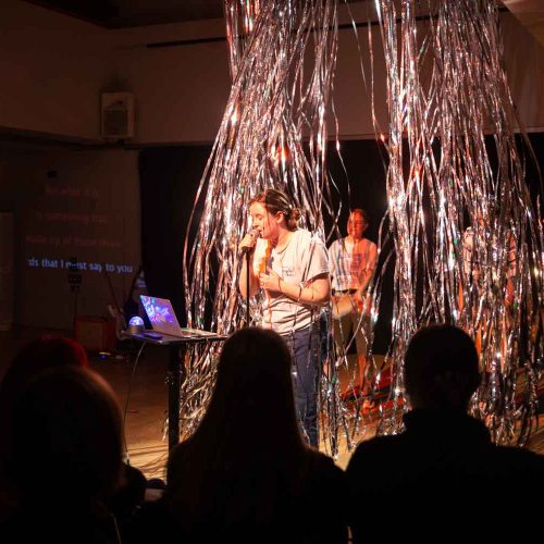 A women sings into a microphone while looking at a laptop. Behind her is glittering strings floating down from the ceiling with people blowing the strings behind her.