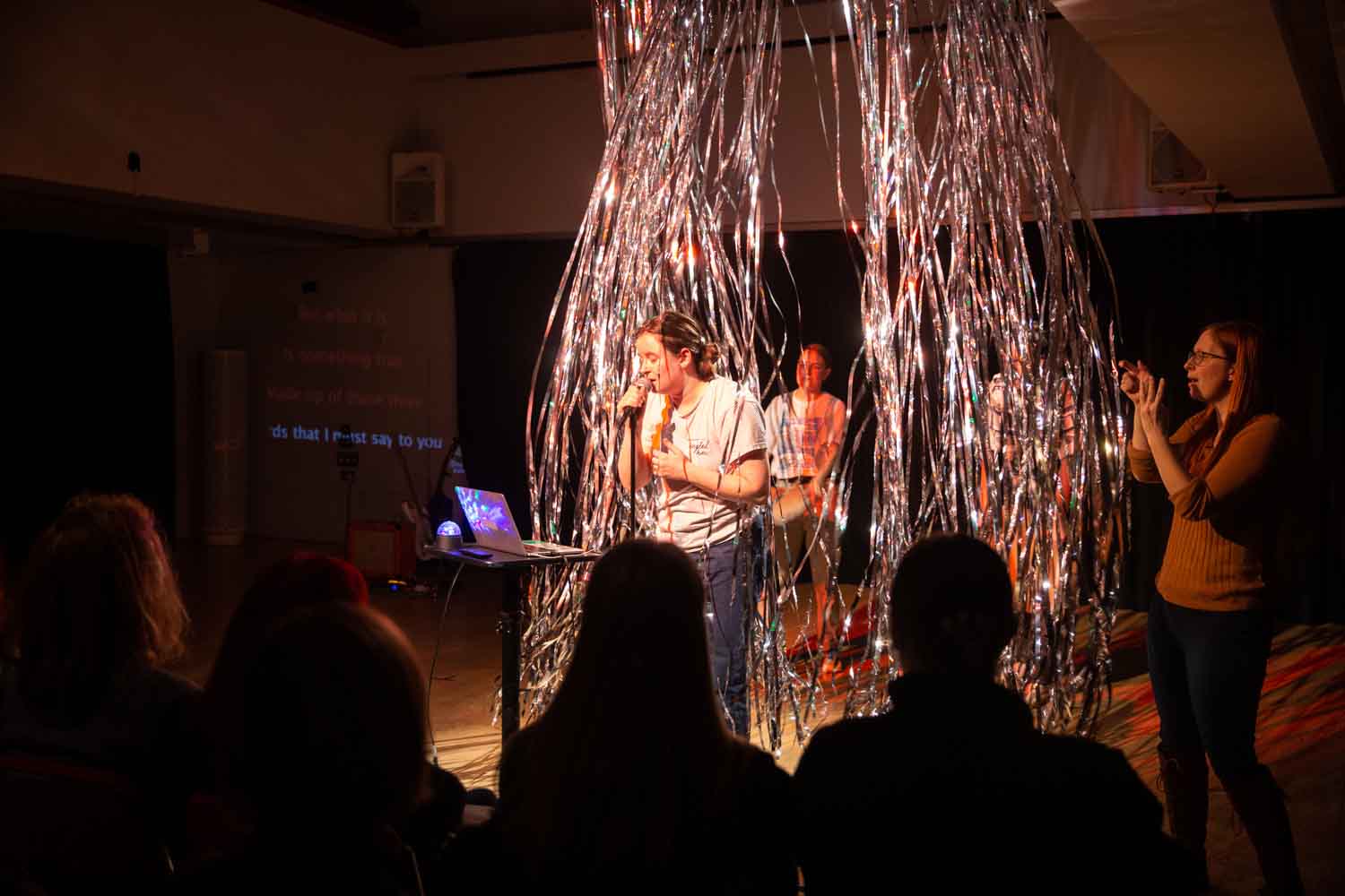 A women sings into a microphone while looking at a laptop. Behind her is glittering strings floating down from the ceiling with people blowing the strings behind her.