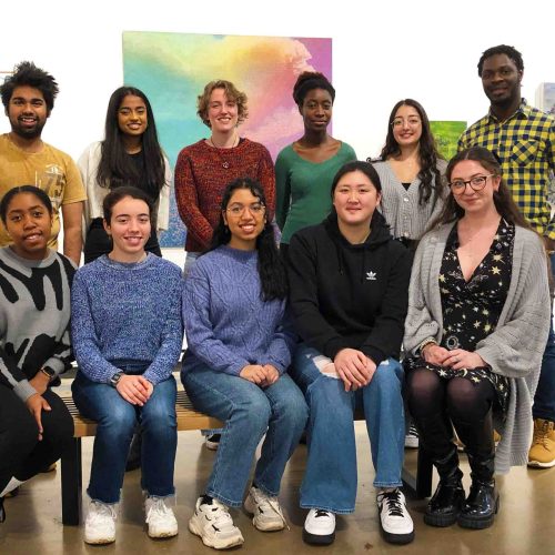 A group of students, front row sitting back row standing, smiling at a camera in a art gallery.