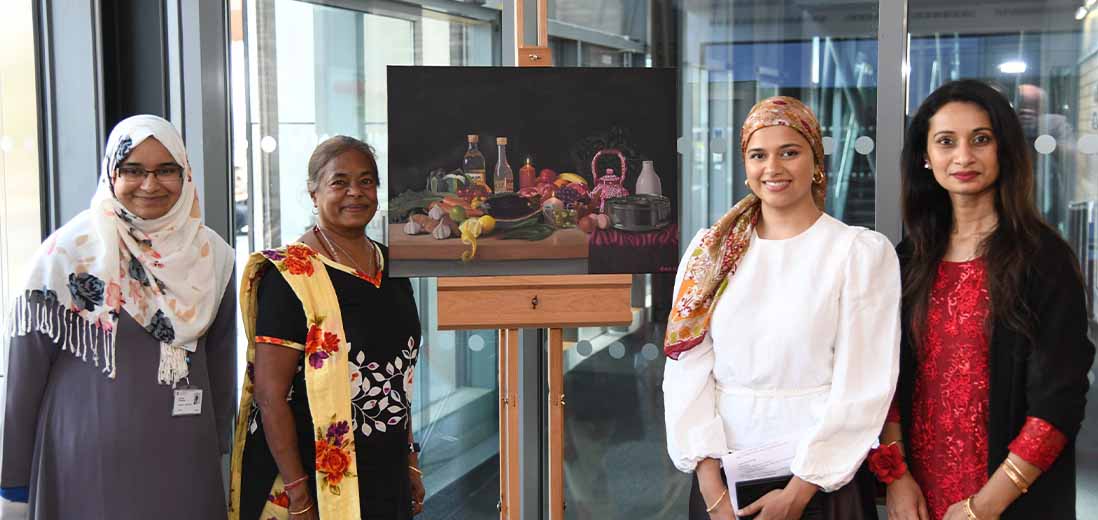 Four women smiling with a oil painting of a still life in the middle of them on an easel.