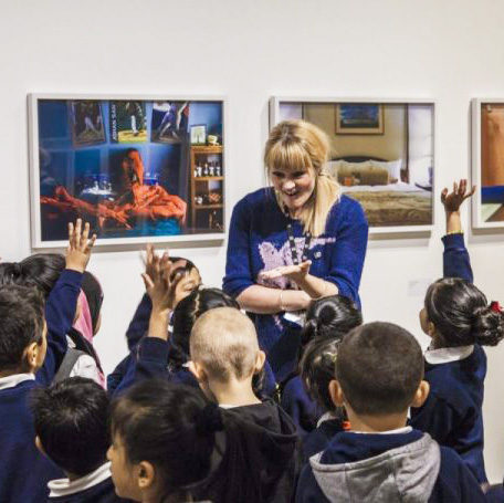 A women stood in front of a large group primary school children as they discuss the artwork behind them.