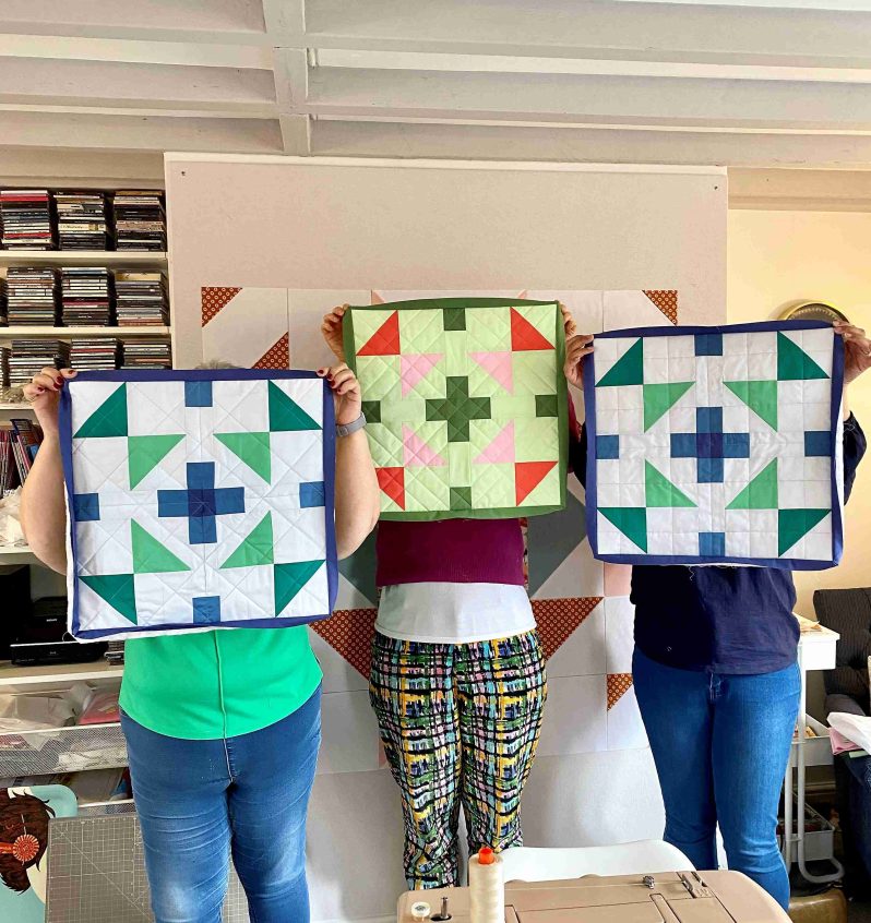 Three people holding up fabric geometric quilts they are holding in front of their faces in a studio.