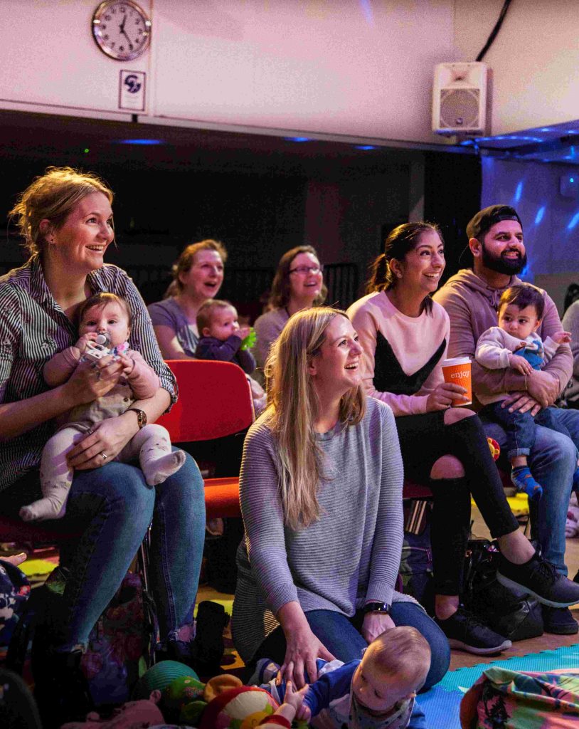 A group of families and babies sat in a theatre while laughing and smiling.