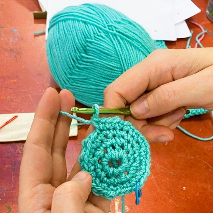 Two hands using a crochet hook to create a circle out of teal yarn.