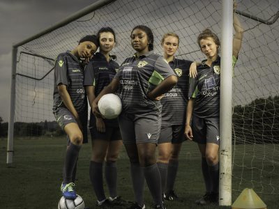 A group of women footballers stood in a pitch with the goal, on a dark and stormy day.