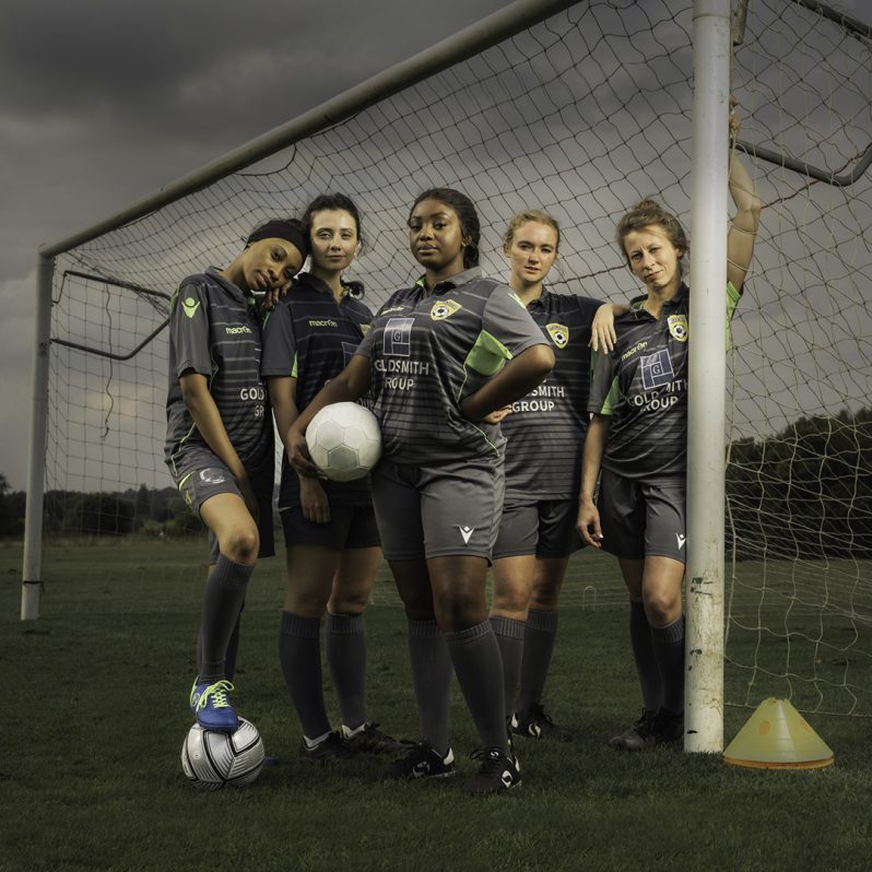 A group of women footballers stood in a pitch with the goal, on a dark and stormy day.