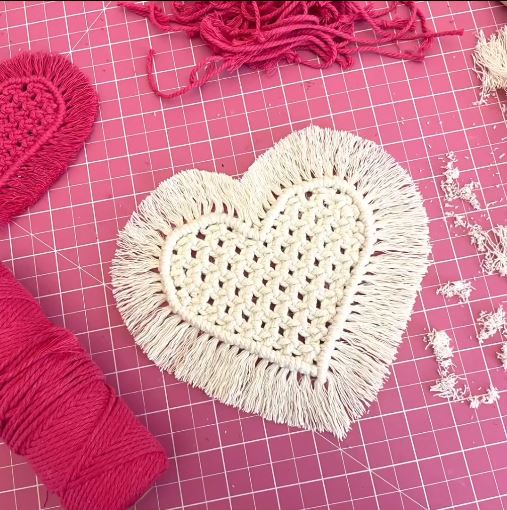 A macramé heart with fringes sitting on top of a pink tiled table next to pink yarn.