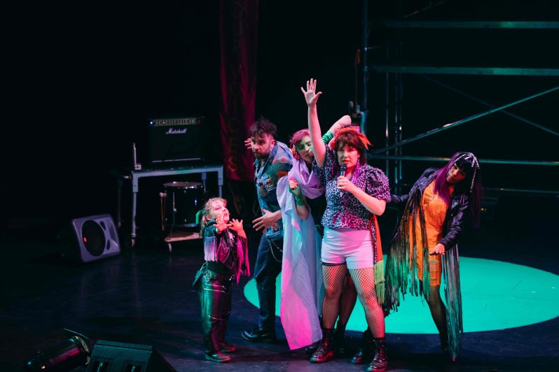 A group of people dressed like colourful punks on a dark stage, dancing together and singing to an audience.