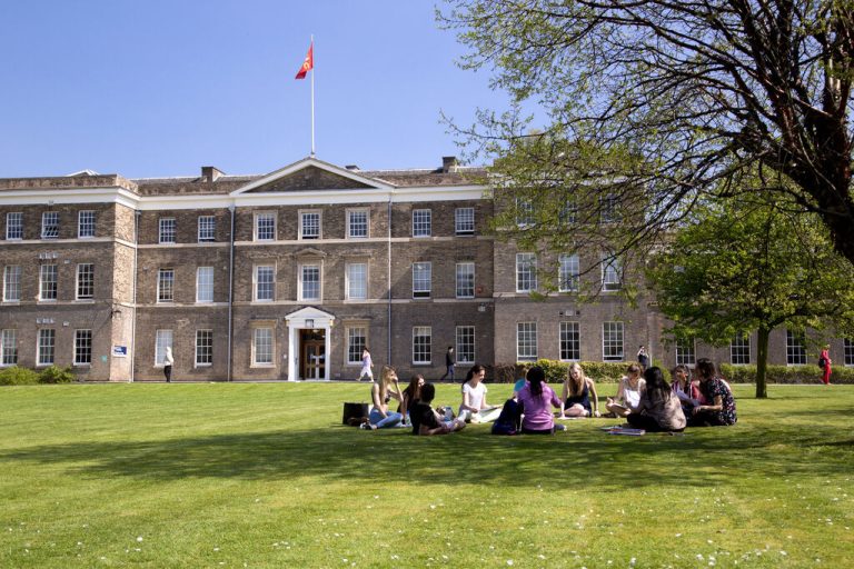 A large, old university building in the background with blue skies. In the foreground are a group of people sat in a circle on grass.
