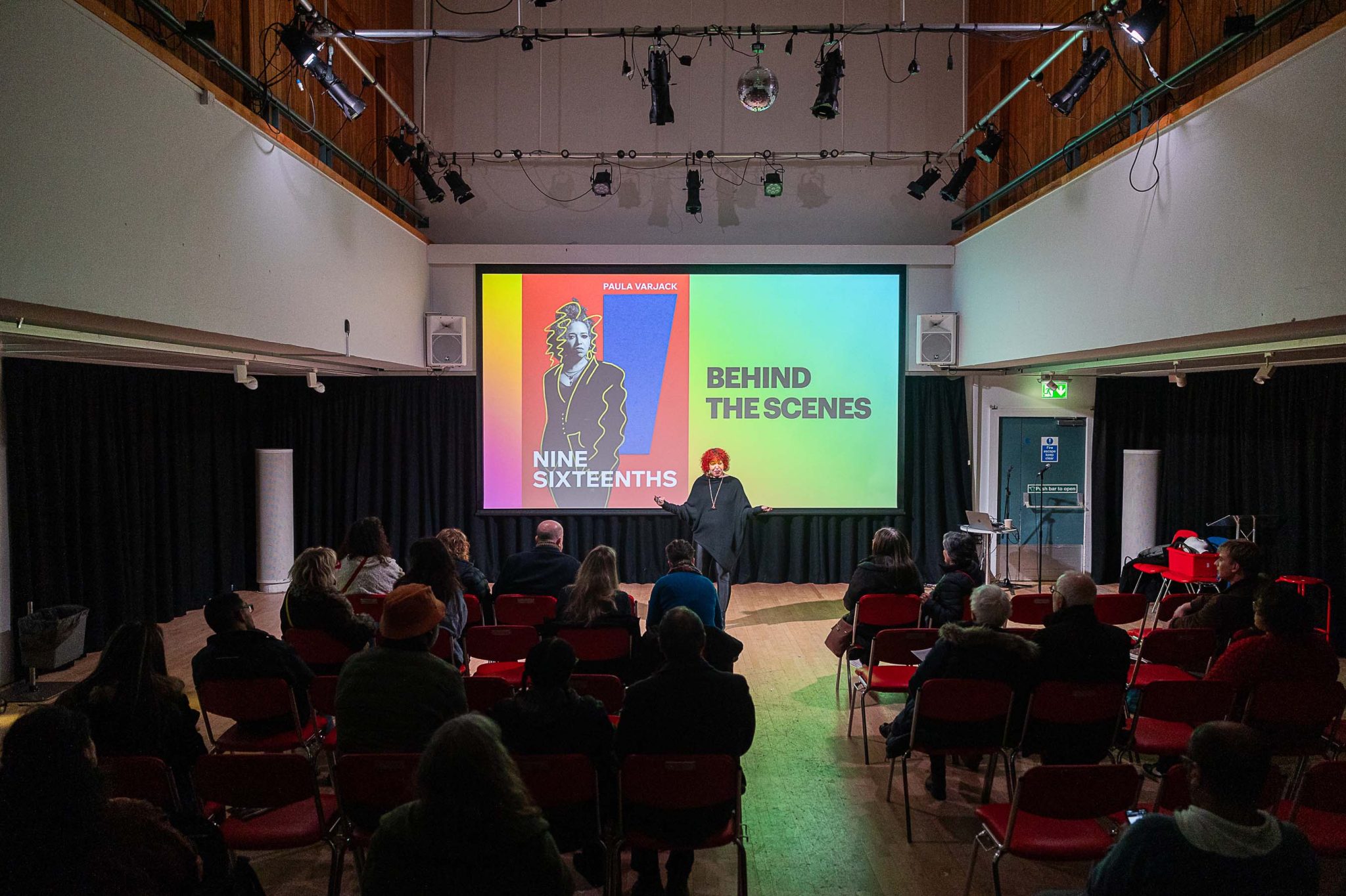 A hall with a large projector, as a women speaks at the front to a crowd.
