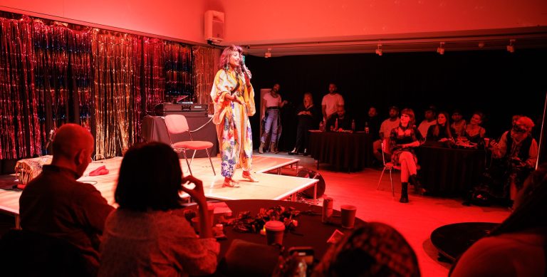 A women dressed in colourful layers, stood an a stage surrounded by an audience. The room is bathed in red lighting.