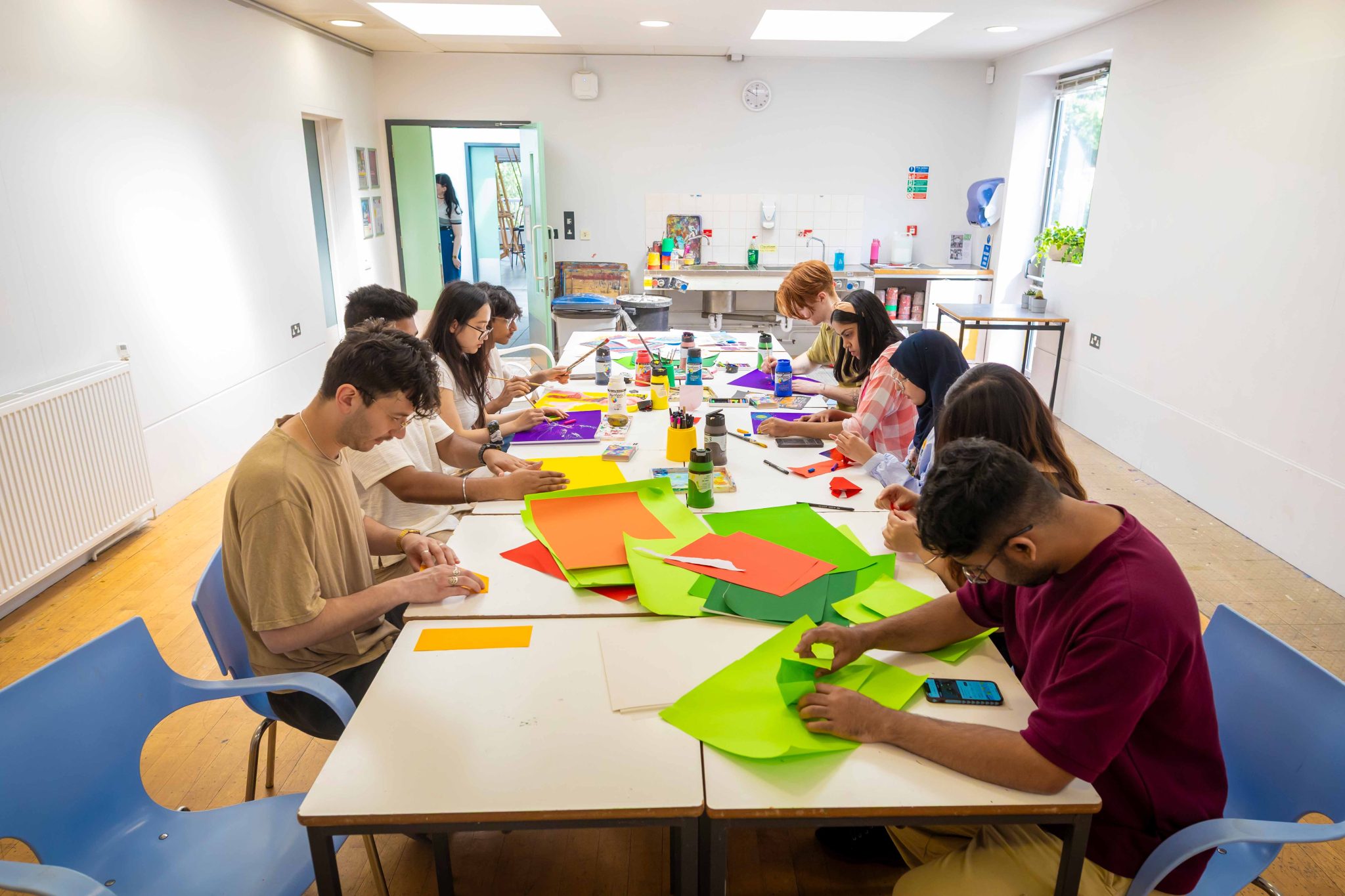 A studio with tables covered in art materials. Students are sat around the tables creating.