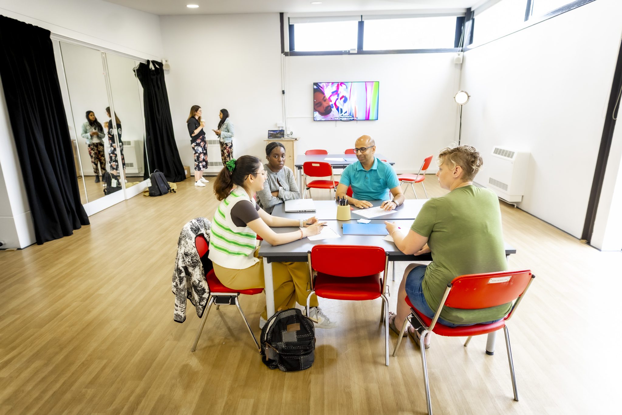 A group of people sat in a room around a table in a studio with mirrors to the left and a tv in the background.