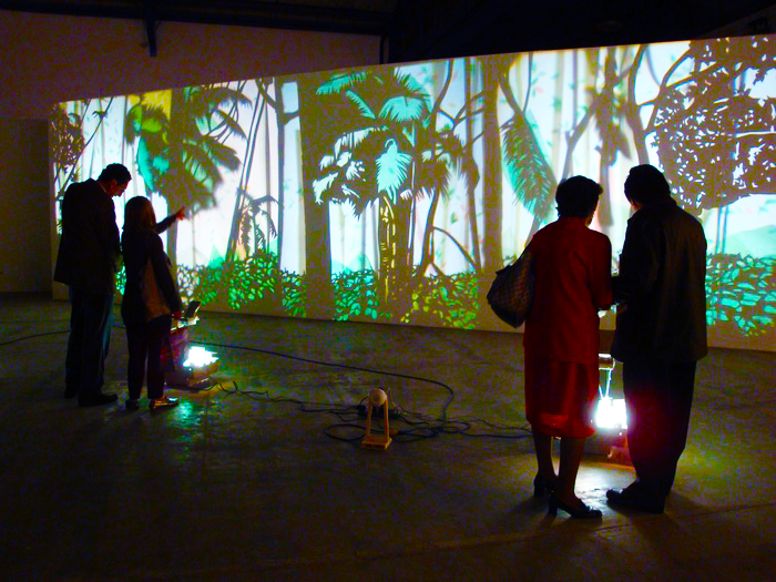 People in a dark room looking at a wall projection of a colourful jungle scene.