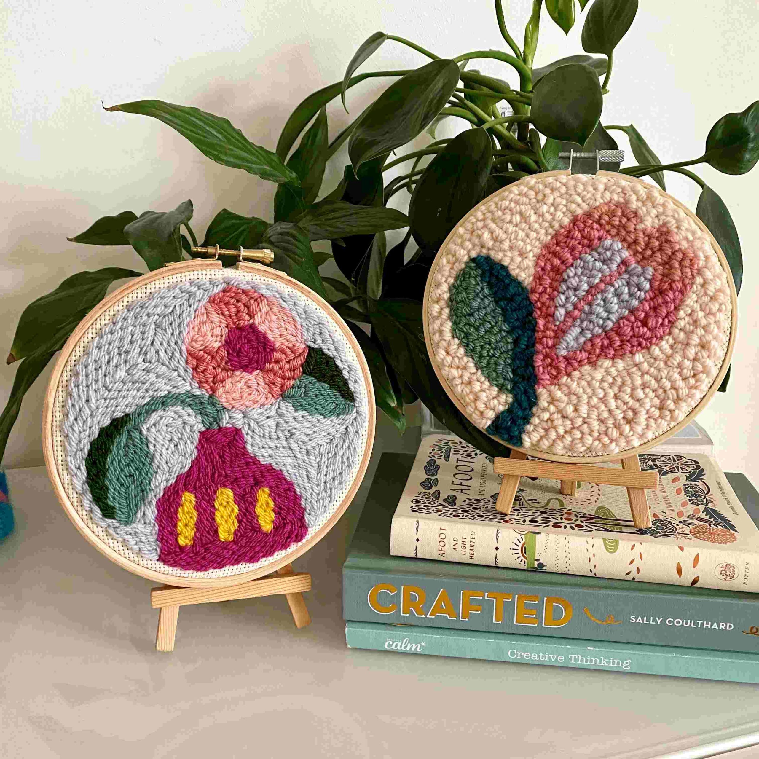 Two punch needle scenes of flowers in wooded hops displayed on top of books.