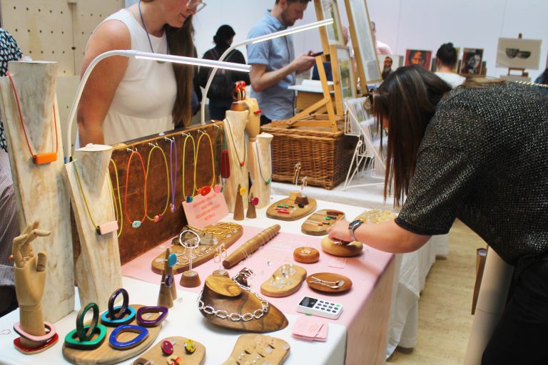 Woman bent over a table looking at jewellery displayed in various ways, in the middle of a craft fair.