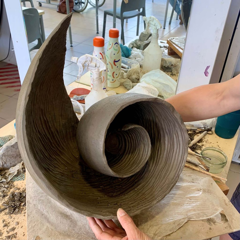 A large textured swirl made out of clay being supported by two hands in an art studio.