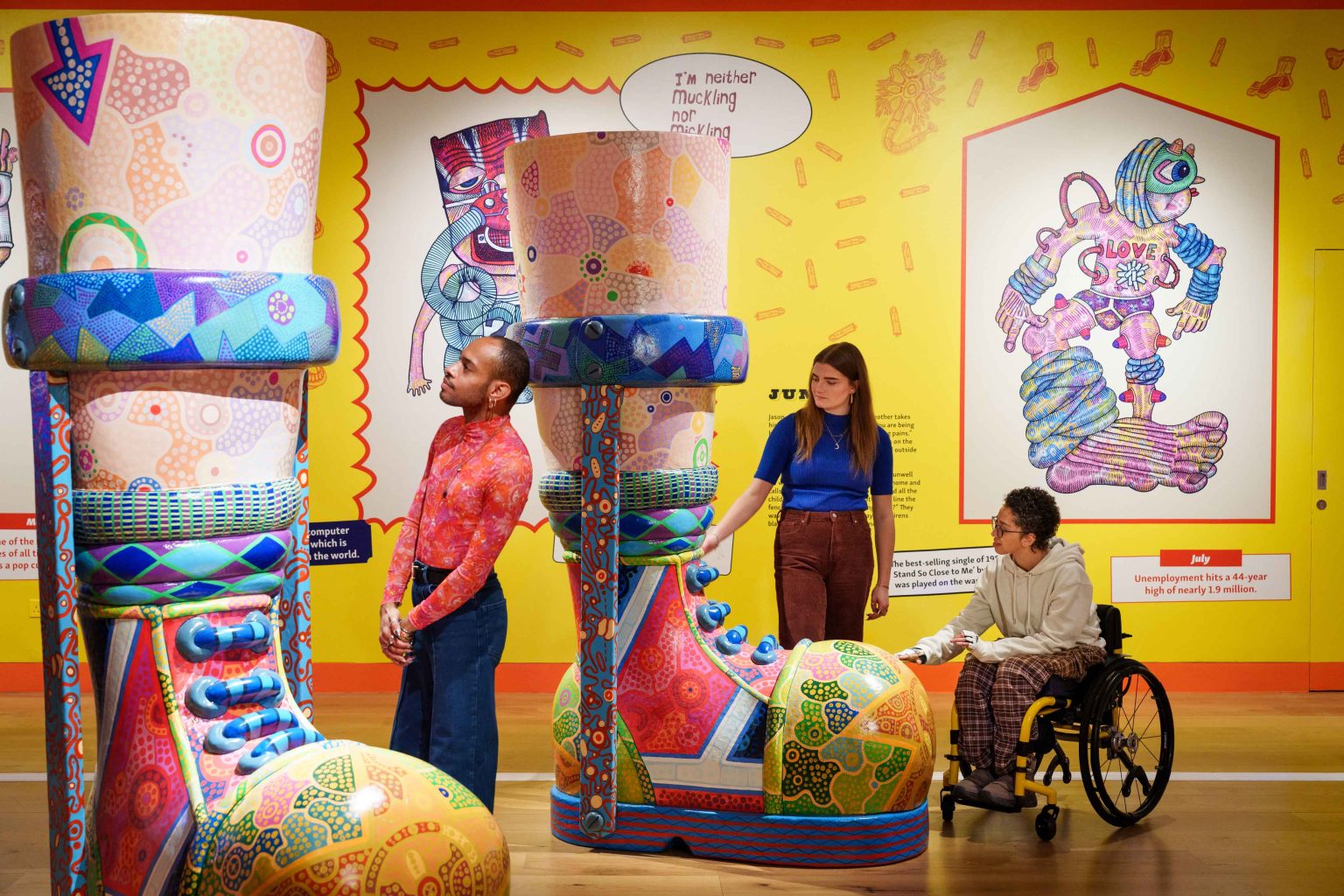 Three people, two standing and one in a wheelchair, looking at a giant pair of cartoonish, colourful legs with braces and colourful shoes. Behind them is a yellow wall with giant drawings of stylised people.
