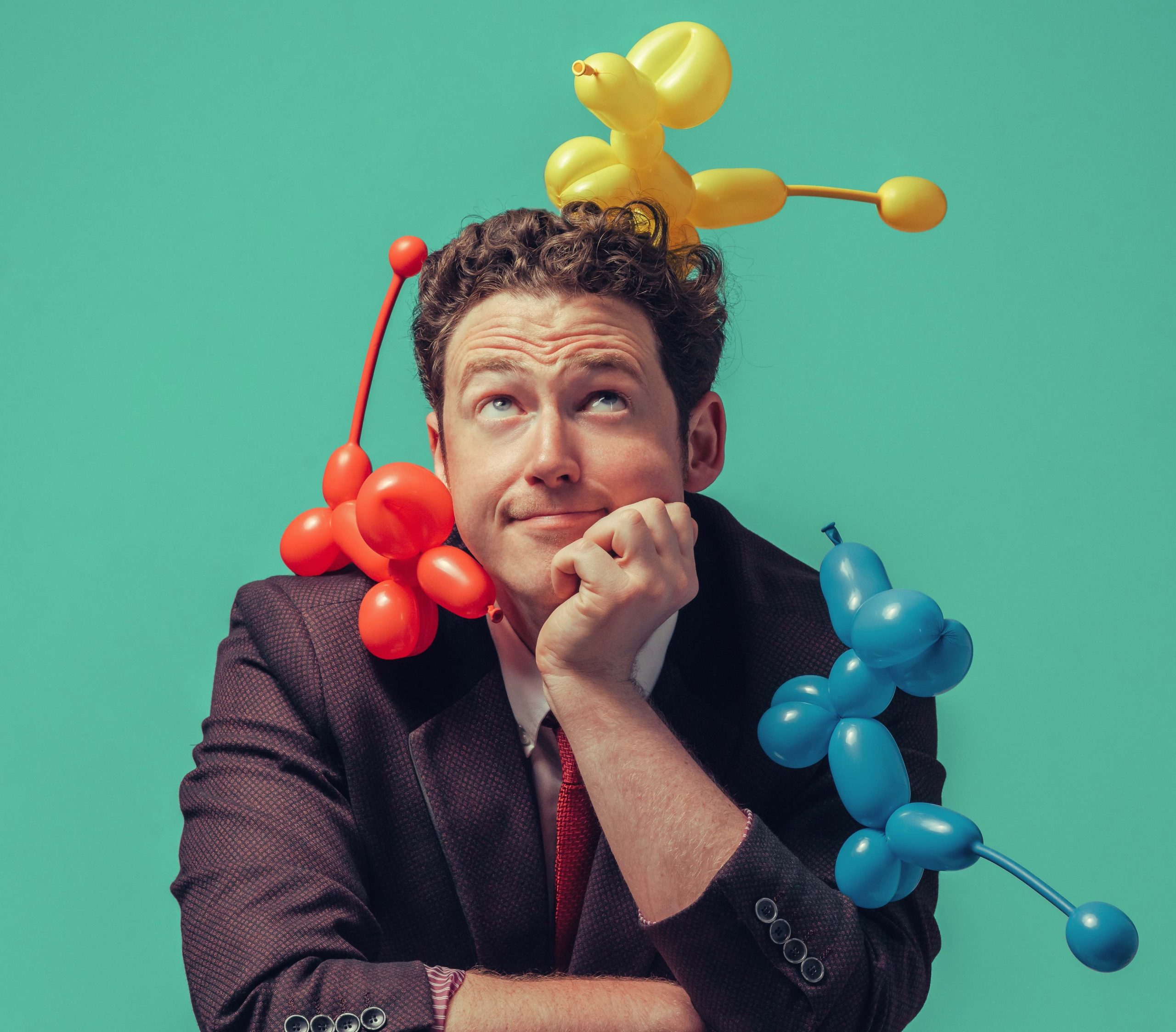 A white man in a suit surrounded by balloon animals looking up happily.