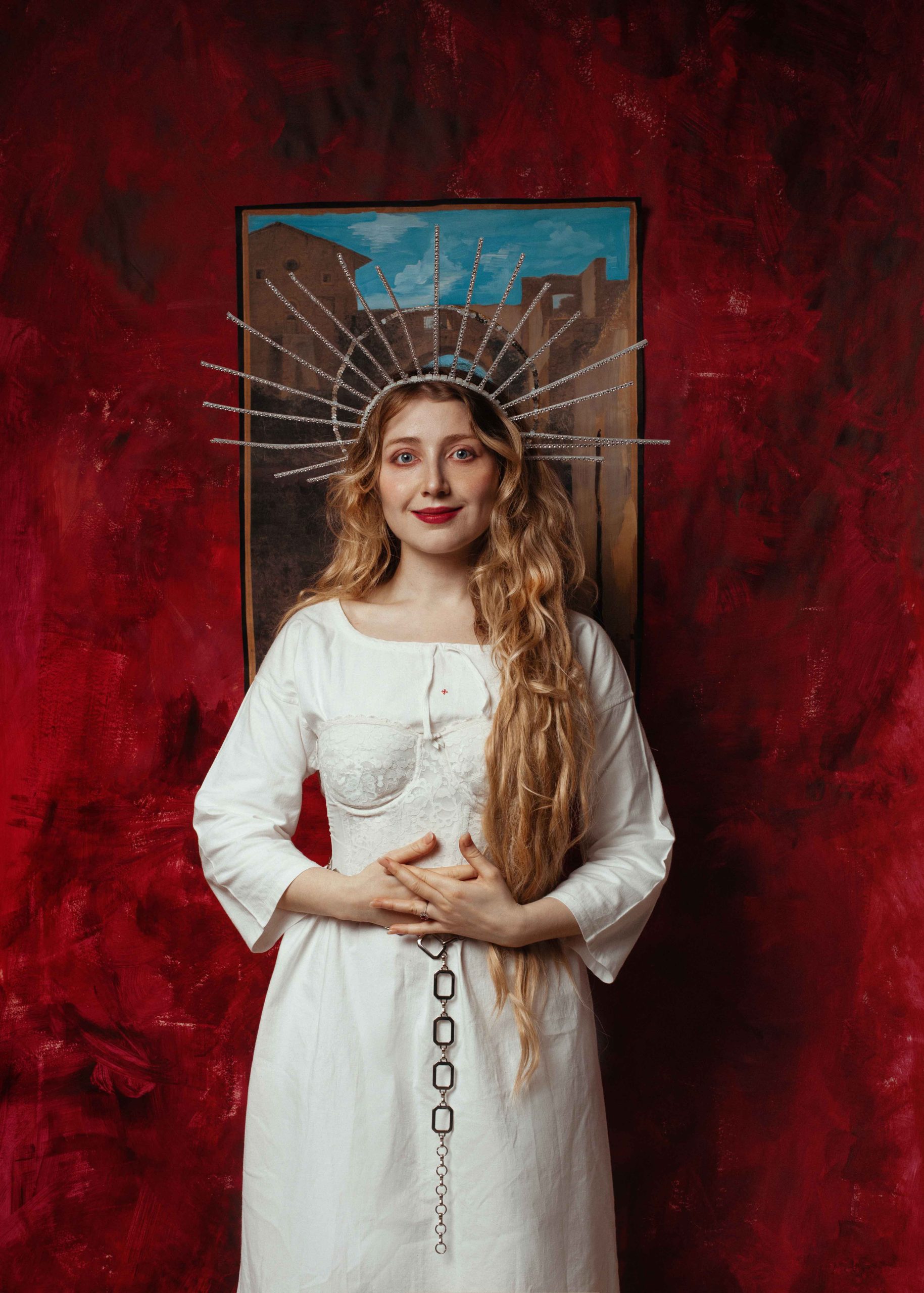 A white women with long blonde hair dressed in a white dress and gold halo, standing in front of a painting of buildings and a red velvet wall.