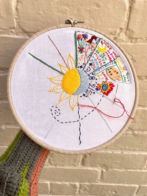 An embroidery hoop with an embroidery of the moon and sun surrounded by different coloured shapes.