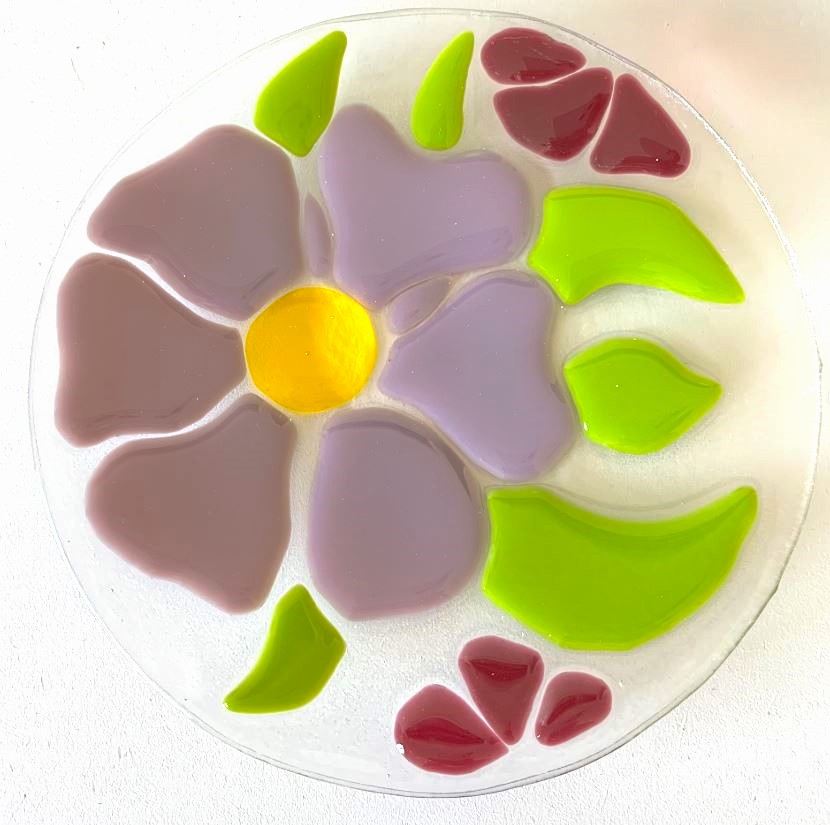 A circle of clear glass with inserts of a glass purple flower with green leaves and red smaller flowers surrounding it.