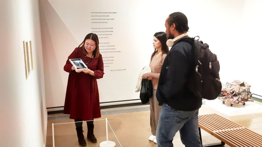 Two people being led on an art gallery tour, looking at art work and their guide holding an iPad.