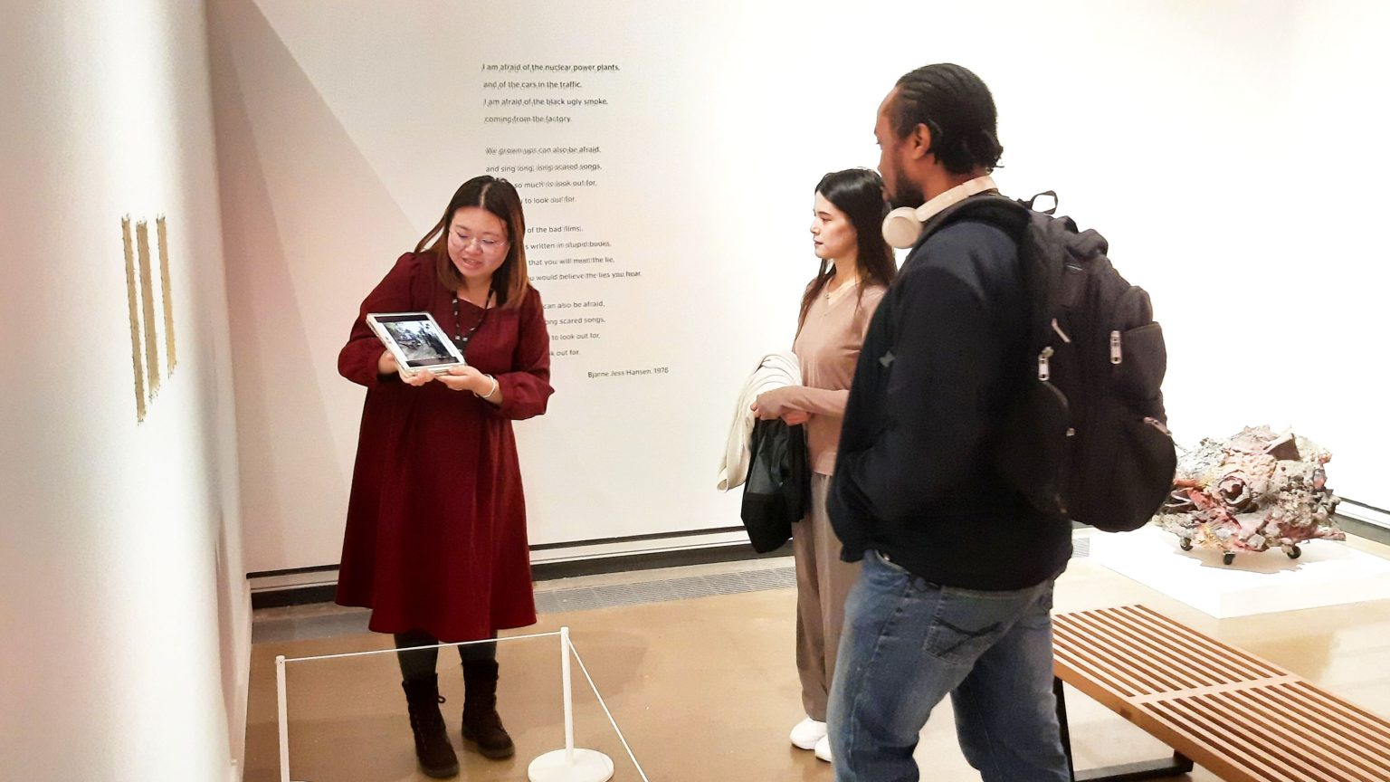 Two people being led on an art gallery tour, looking at art work and their guide holding an iPad.