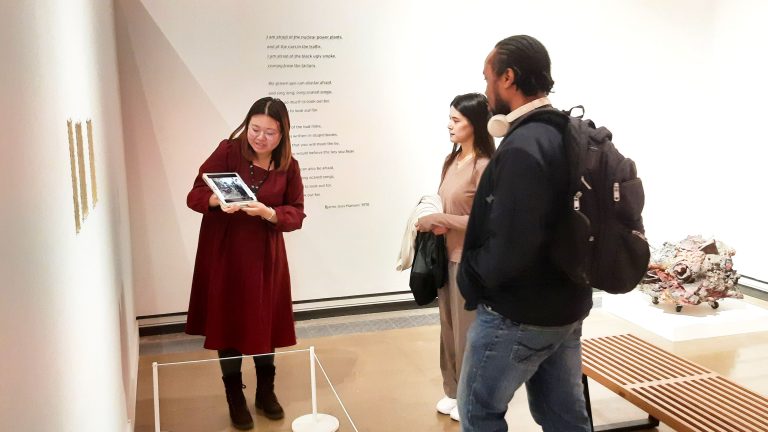 Two people being led on an art gallery tour, looking at art work and their guide holding an iPad.