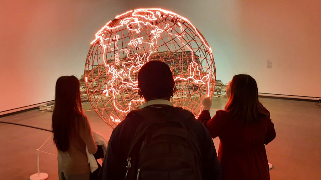 Three people in darkness looking at a giant, red glowing globe in front of them.