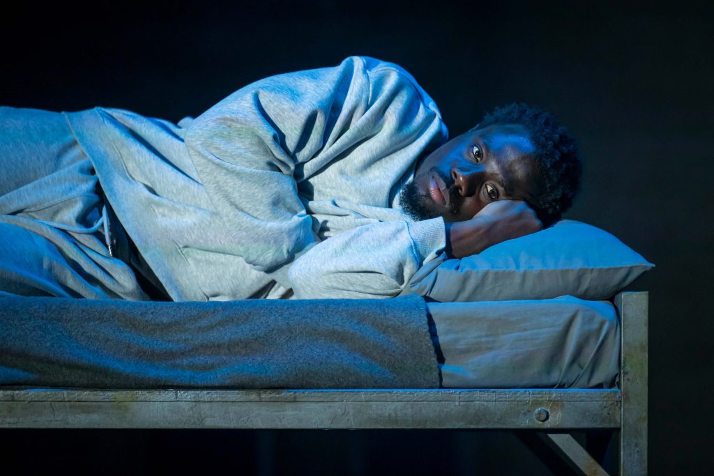A Black man dressed in white, laying down in a dark bed looking unhappy.