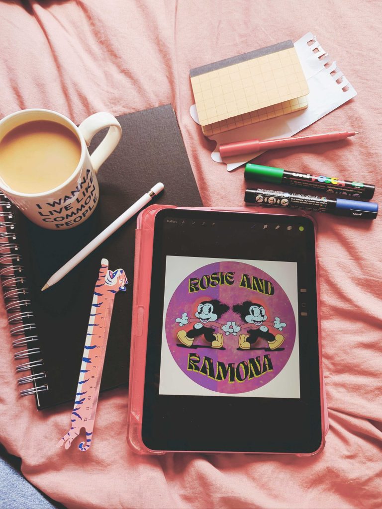 A tablet showing a piece of artwork, surrounded by pens, coffee, notebooks and colours.